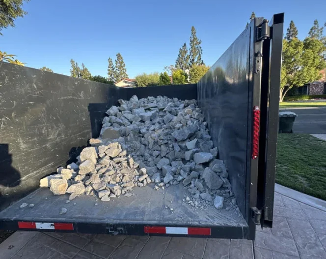 A black dump trailer loaded with grey rock debris, parked on a textured gray driveway under a clear sky.